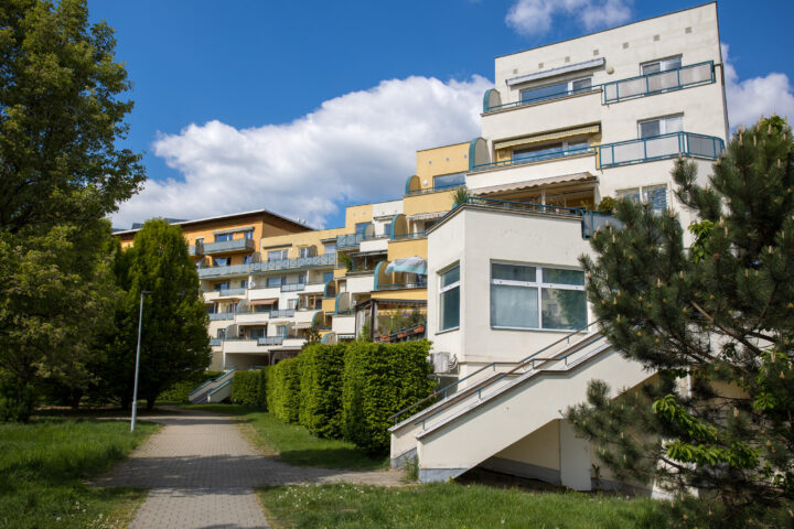 A multi-storey terraced flat complex with light-coloured walls and tiered balconies stands under a bright blue sky. A paved walkway leads through a landscaped area with manicured hedges and trees in the foreground.