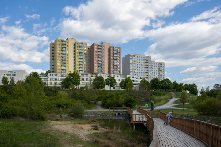 Three tall block of flats with yellow, brown, and grey facades rise above a landscaped park. In the foreground, a wooden footbridge curves over a small pond, where a couple stands looking at the water. Green trees and rolling hills surround the buildings under a blue sky.