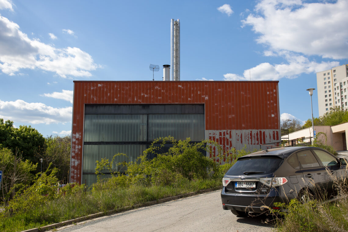 A dark grey Subaru hatchback is parked on a dirt road in front of an industrial building with weathered red corrugated metal walls and tall frosted-glass windows. Two metal chimneys extend from the roof against a bright blue sky with scattered clouds.