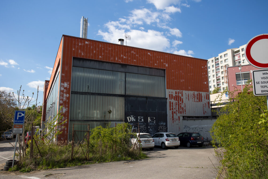 A weathered, orange-panelled industrial building stands under a bright blue sky, with three cars parked in front of its large sliding doors covered in graffiti. Overgrown bushes and a parking sign labelled ZONA E occupy the foreground, with a block of flats visible in the distance.