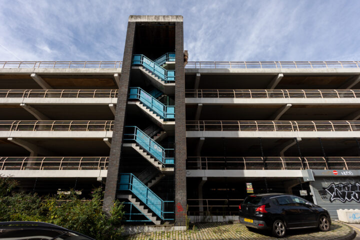 A multi-storey concrete car park features a central dark brick stairwell with bright blue metal stairs. A black hatchback is parked at the base, and graffiti marks the wall near a closed metal shutter. The sky above is bright with wispy clouds.
