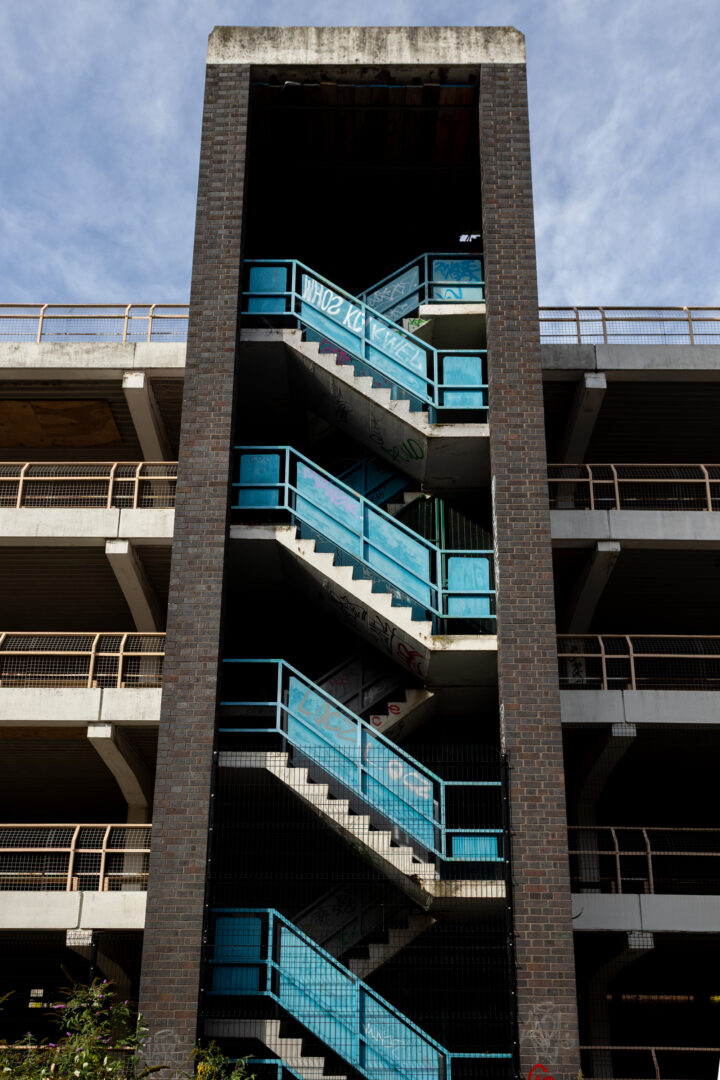 A vertical brick stairwell tower stands between levels of an open-air concrete car park. Bright blue railings follow the zig-zagging stairs, which are marked by layers of graffiti. A thin mesh fence partially covers the ground level, and a cloudy blue sky sits above.